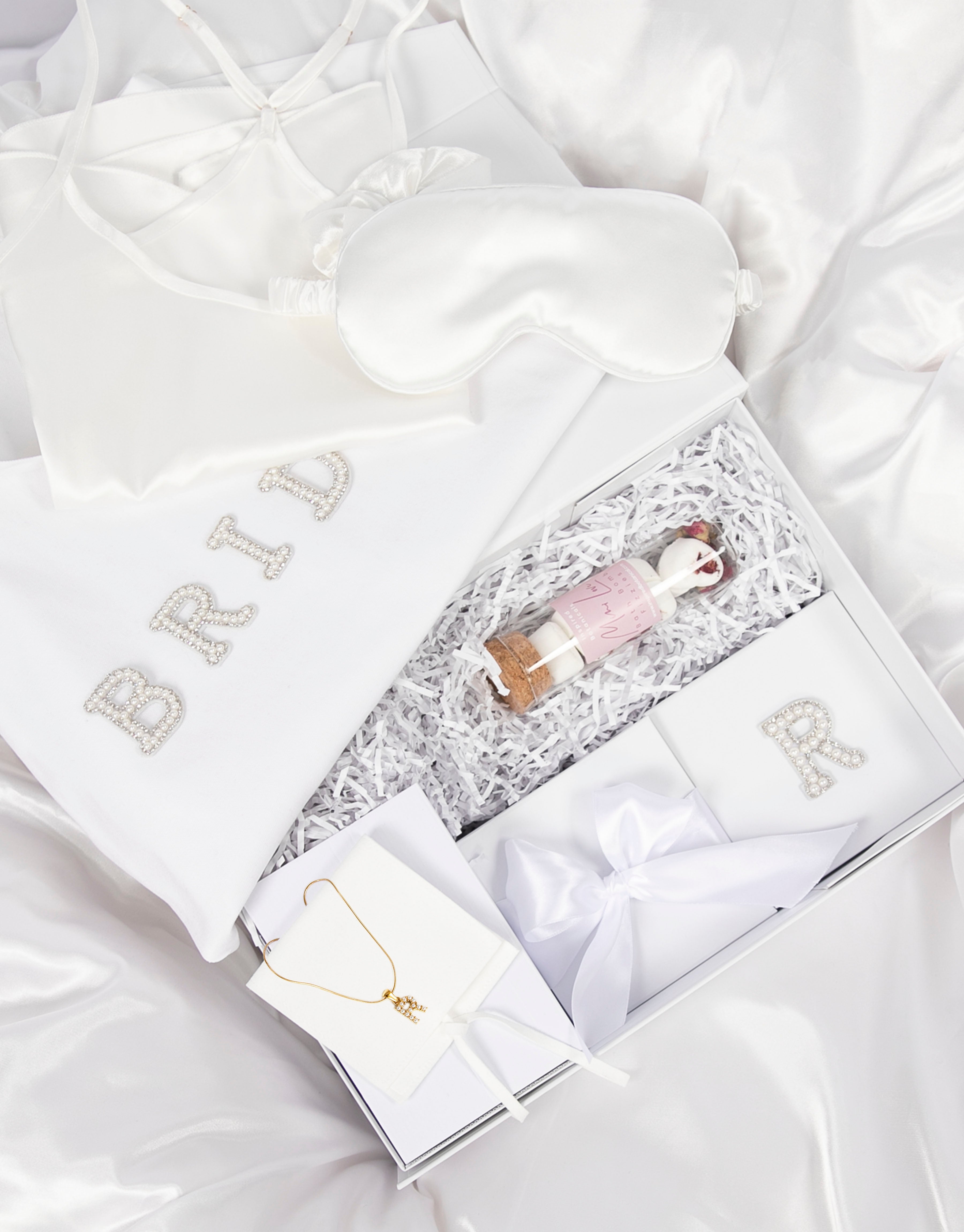 23Dreams Whitehaven Bliss Bridal Box: white tote bag with "bride" pearl and rhinestone patches, 2 petite white gift boxes; one Mini Love hearts Bath Bomb Fizzies; hamptons deluxe candle whitehaven sea salt; ultra soft satin sleep eye mask white; scrunchie soft silky satin white; luxe silky camisole set white; Elegant 18K Gold Plated Letter Pearl Necklace; glamour pearls hair pin set in gold 18 pins; white pearls gold hair pin set luxe glam and white pearled hair clip set in gold timeless elegance.
