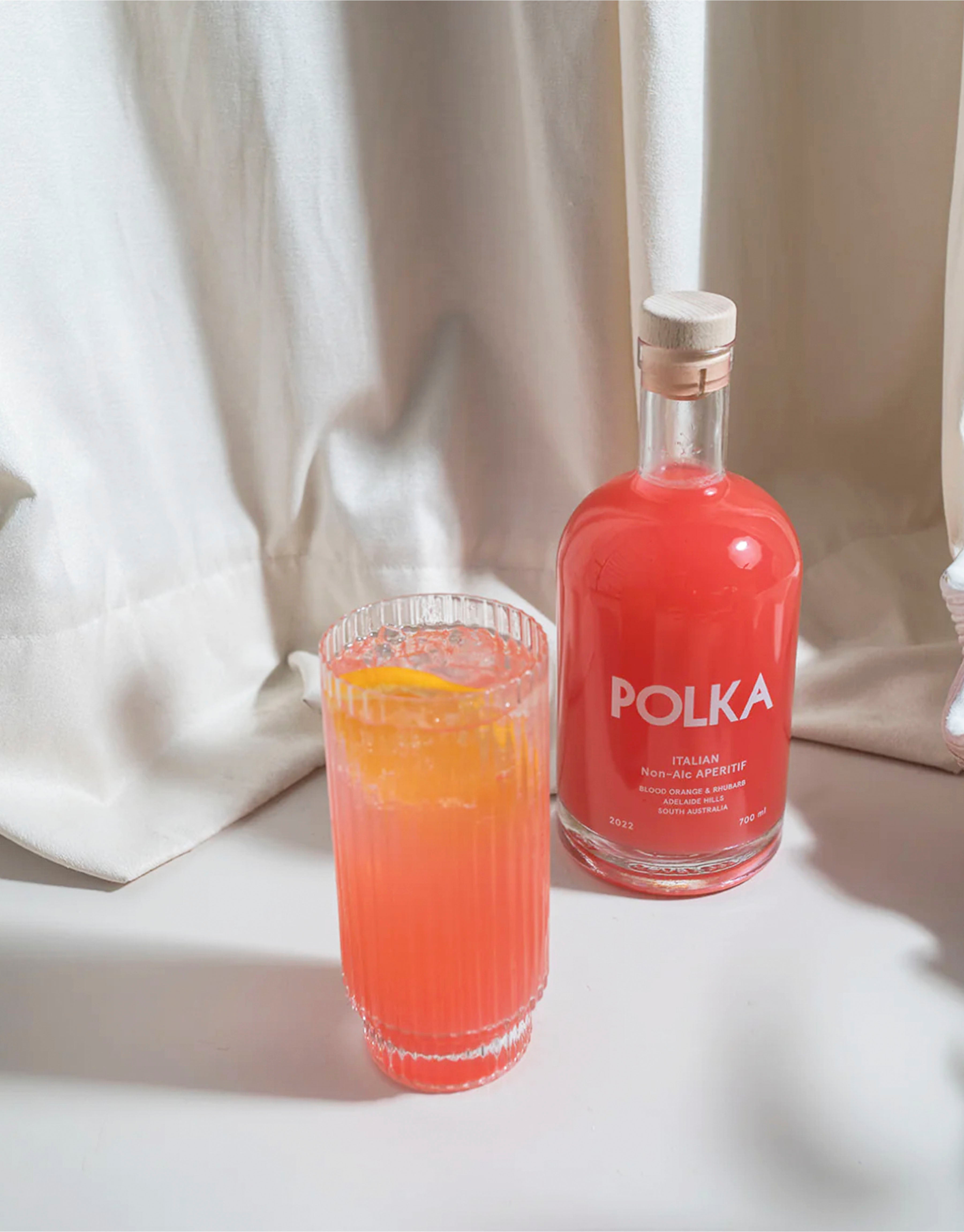 Un-opened bottle of non-alcoholic Italian aperitif next to a tall 3glass of Aperitif garnished with a slice of orange.