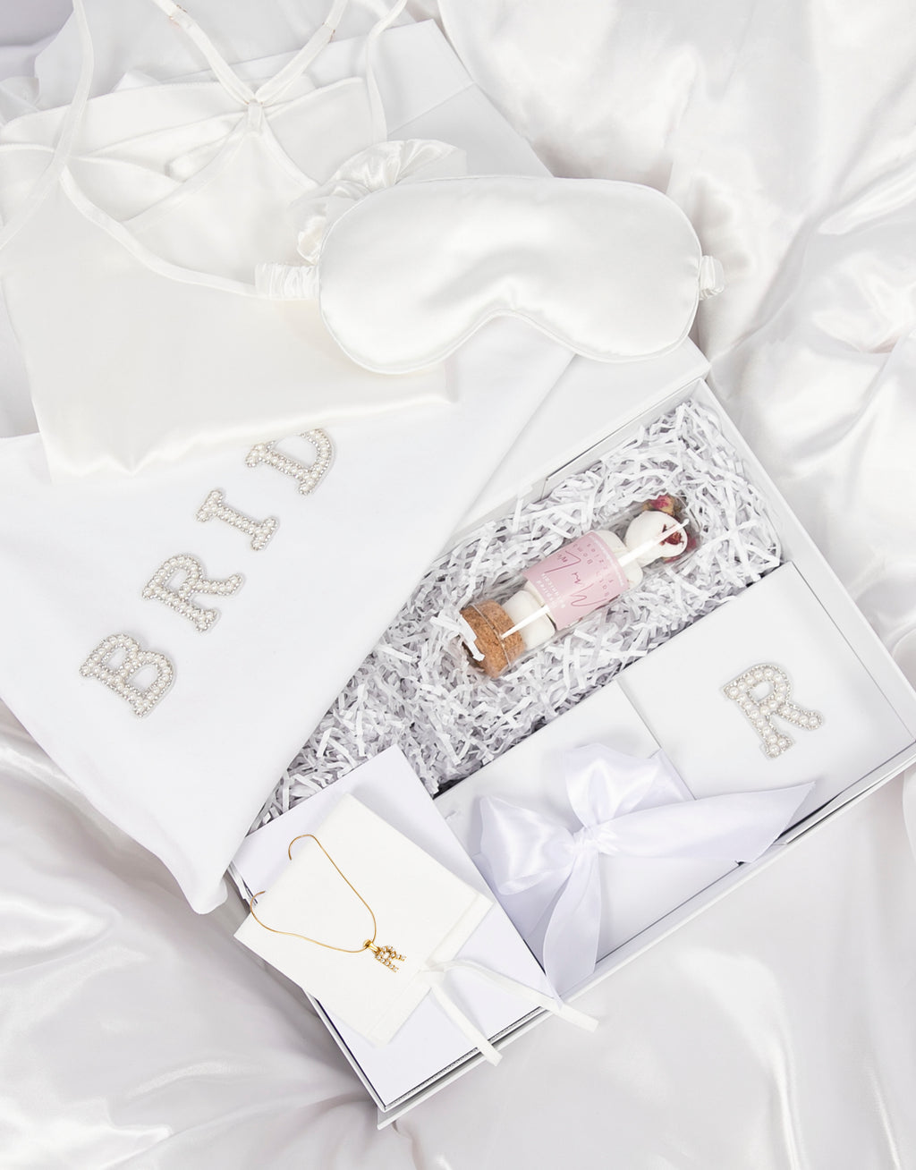 23Dreams Whitehaven Bliss Bridal Box: white tote bag with "bride" pearl and rhinestone patches, 2 petite white gift boxes; one Mini Love hearts Bath Bomb Fizzies; hamptons deluxe candle whitehaven sea salt; ultra soft satin sleep eye mask white; scrunchie soft silky satin white; luxe silky camisole set white; Elegant 18K Gold Plated Letter Pearl Necklace; glamour pearls hair pin set in gold 18 pins; white pearls gold hair pin set luxe glam and white pearled hair clip set in gold timeless elegance.